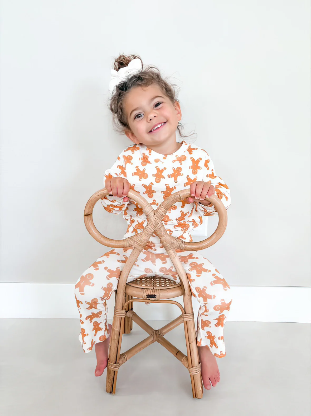 A young girl sits on a wooden chair wearing a two-piece set with a long-sleeved sweatshirt and sweatpants. The fabric design features gingerbread men on a white background.